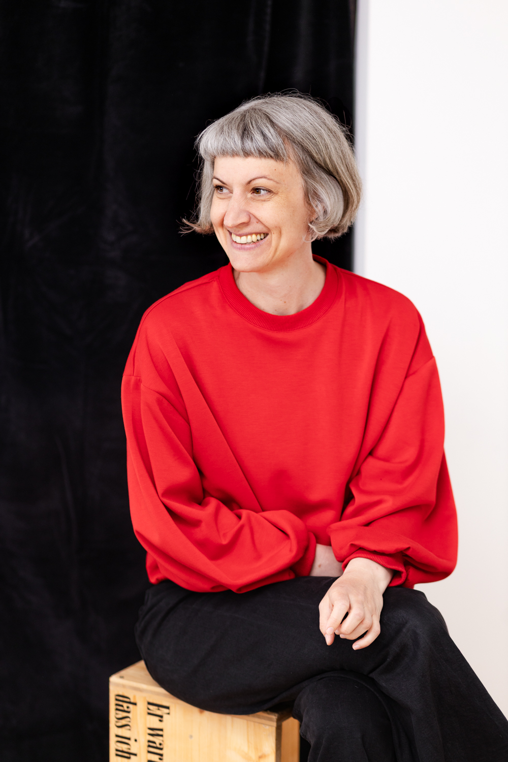 Portraitfotografie einer Kostuembildnerin, Frau sitzt lachend vor halb schwarzem, halb weißem Hintergrund, in ihrem Atelier in Wien